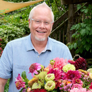 A man with short gray hair and glasses smiles in behind a large bouquet of flowers.