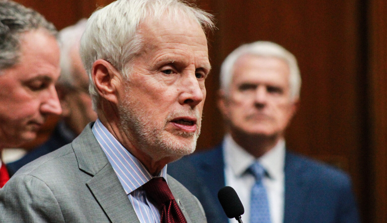 Representative Bob Behning speaks at a microphone. He is a White man with white hair and stubble. He wears a suit with a dark red tie.