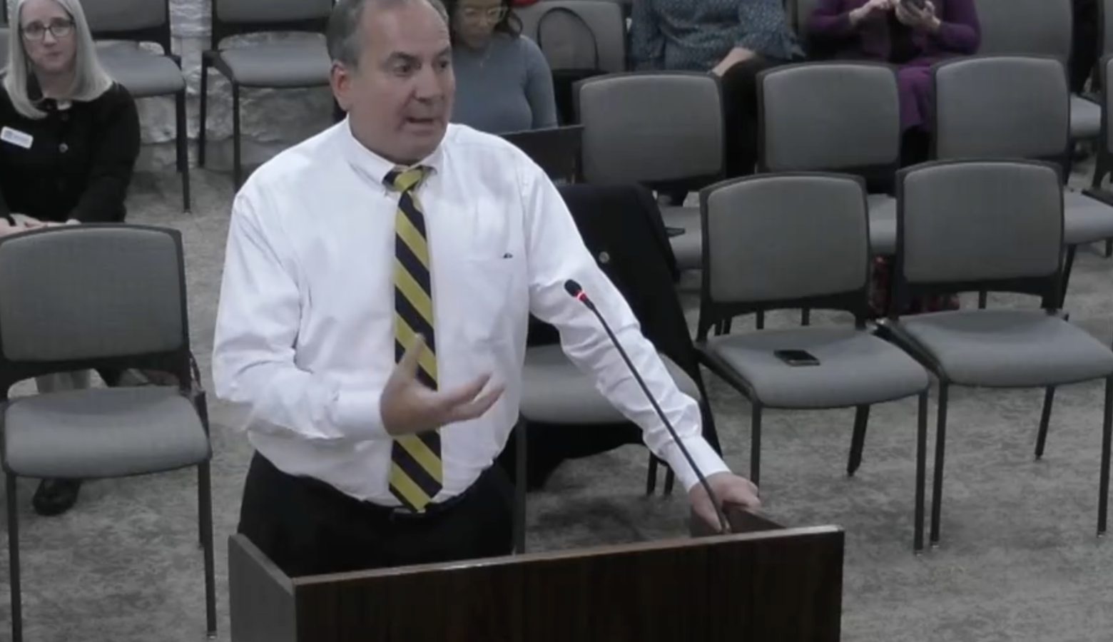 Mike Bohacek stands behind a podium with one hand outstretched. He wears a black and yellow tie with a white button down shirt.