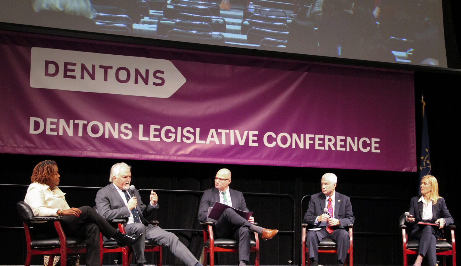 Representatives Robin Shackleford and Brad Barrett and Senators Ed Charbonneau and Shelli Yoder sitting in chairs on a stage, with Barrett speaking into a microphone. The banner behind the lawmakers reads "Dentons Legislative Conference" in white text on a purple background.