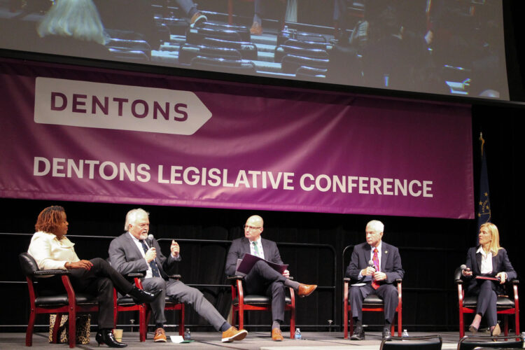 Representatives Robin Shackleford and Brad Barrett and Senators Ed Charbonneau and Shelli Yoder sitting in chairs on a stage, with Barrett speaking into a microphone. The banner behind the lawmakers reads "Dentons Legislative Conference" in white text on a purple background.