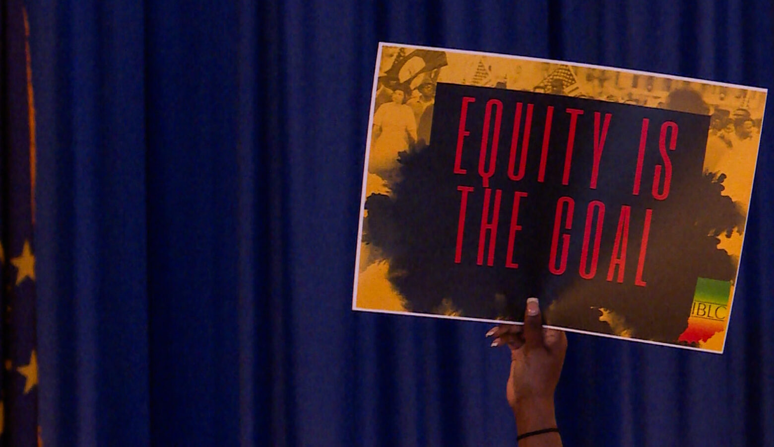 A Black woman holds a sign up above the heads of the crowd that reads "Equity is the goal." To the far left of the sign she's hold is an Indiana state flag.