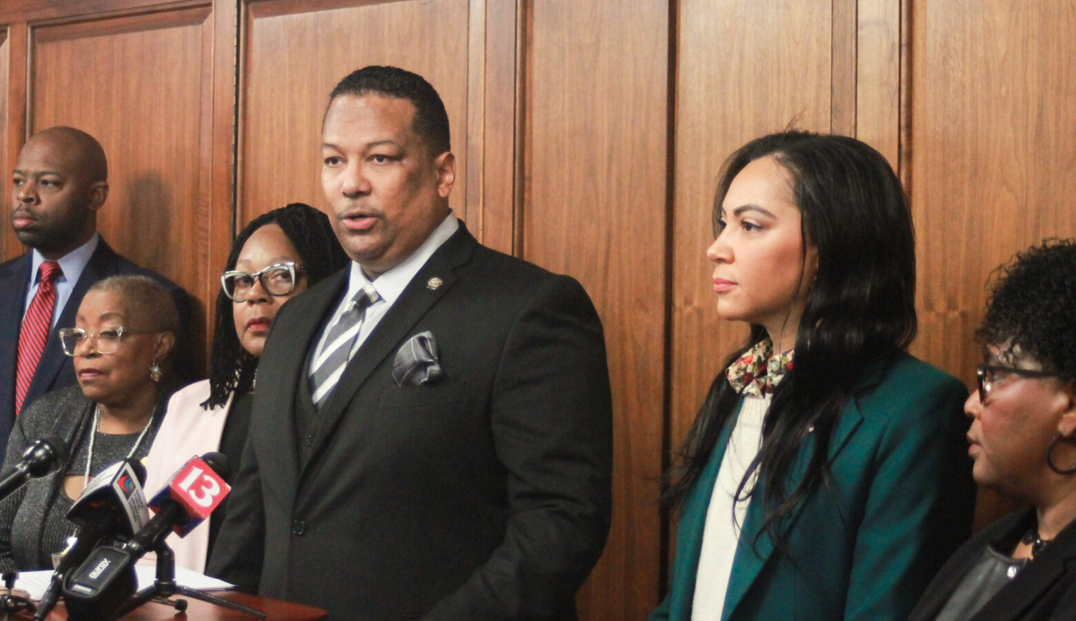 Earl Harris Jr. speaks at a lectern with other members of the Black Caucus standing on either side of him. Harris is a Black man with black hair and goatee. He is wearing a suit and tie.