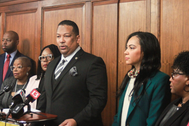 Earl Harris Jr. speaks at a lectern with other members of the Black Caucus standing on either side of him. Harris is a Black man with black hair and goatee. He is wearing a suit and tie.