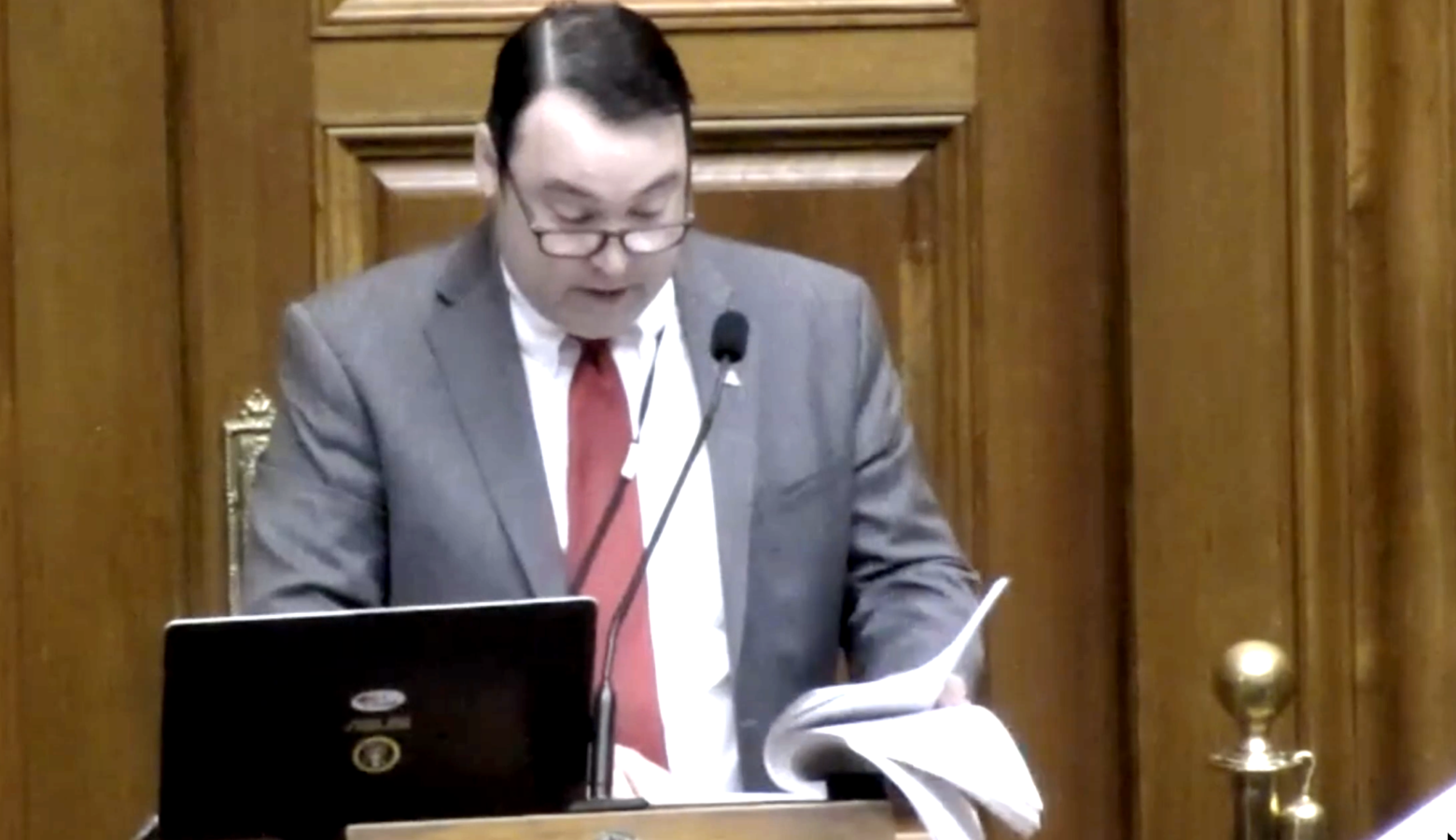 Joel Hand stands behind a podium and looks down at a thick stack of papers while speaking into the microphone. Hand is a White man and he is wearing a red tie and suit jacket.