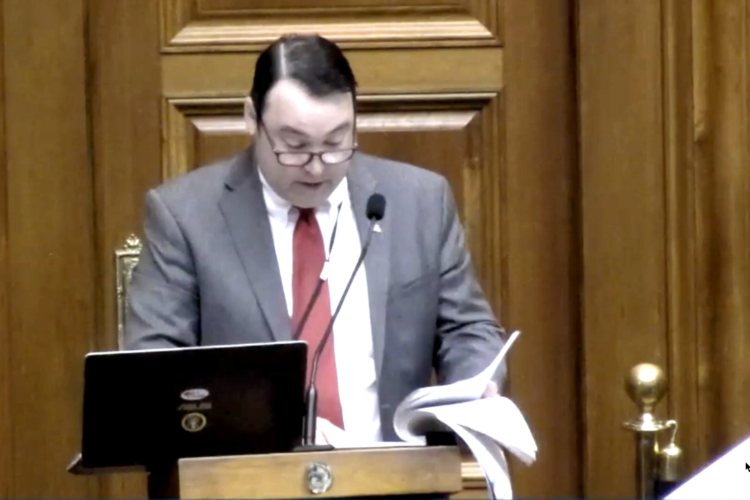 Joel Hand stands behind a podium and looks down at a thick stack of papers while speaking into the microphone. Hand is a White man and he is wearing a red tie and suit jacket.