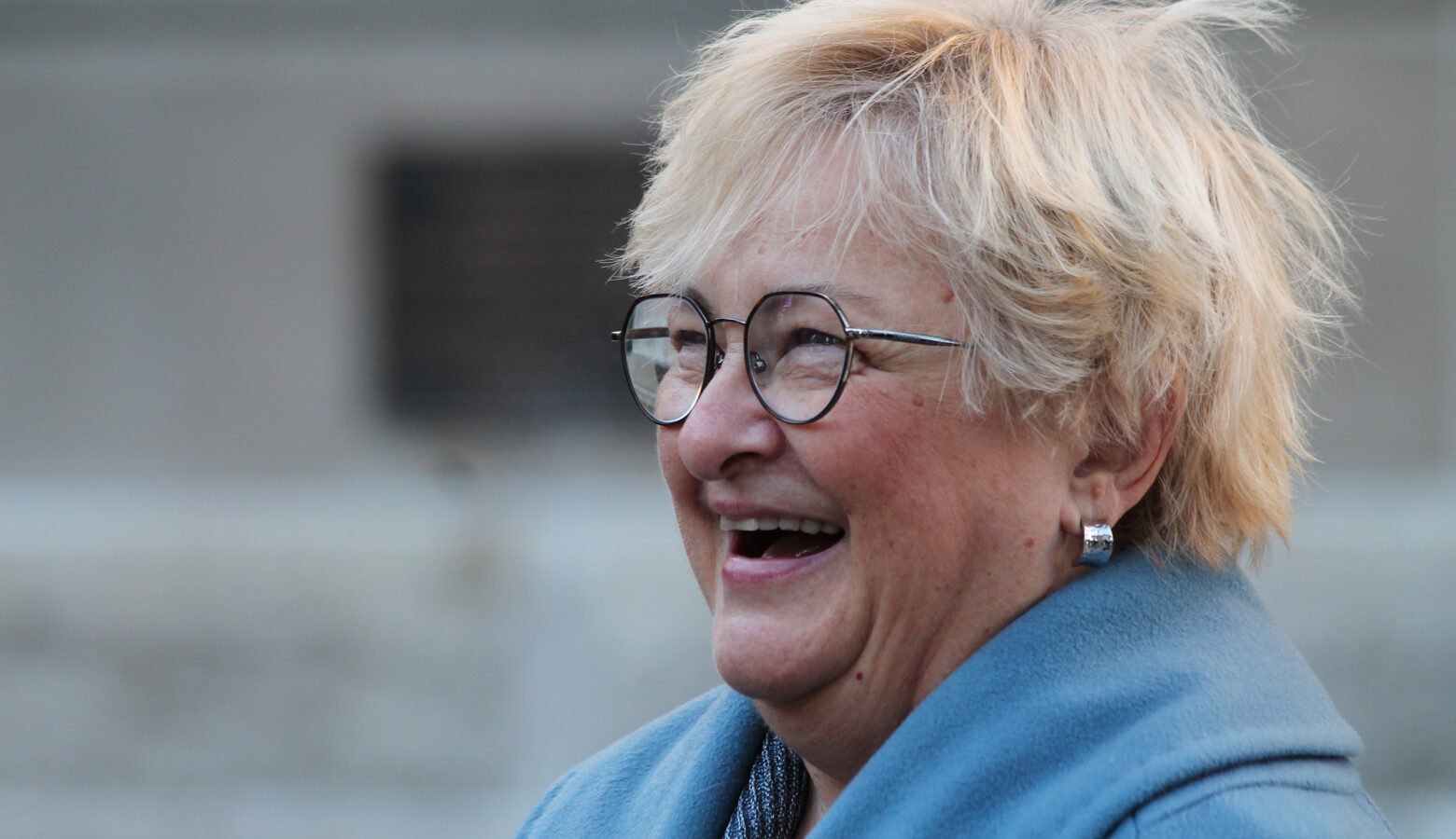 Karen Tallian smiles on the steps of the Indiana Statehouse. Tallian is a White woman with blonde hair. She is wearing glasses and a light blue coat.