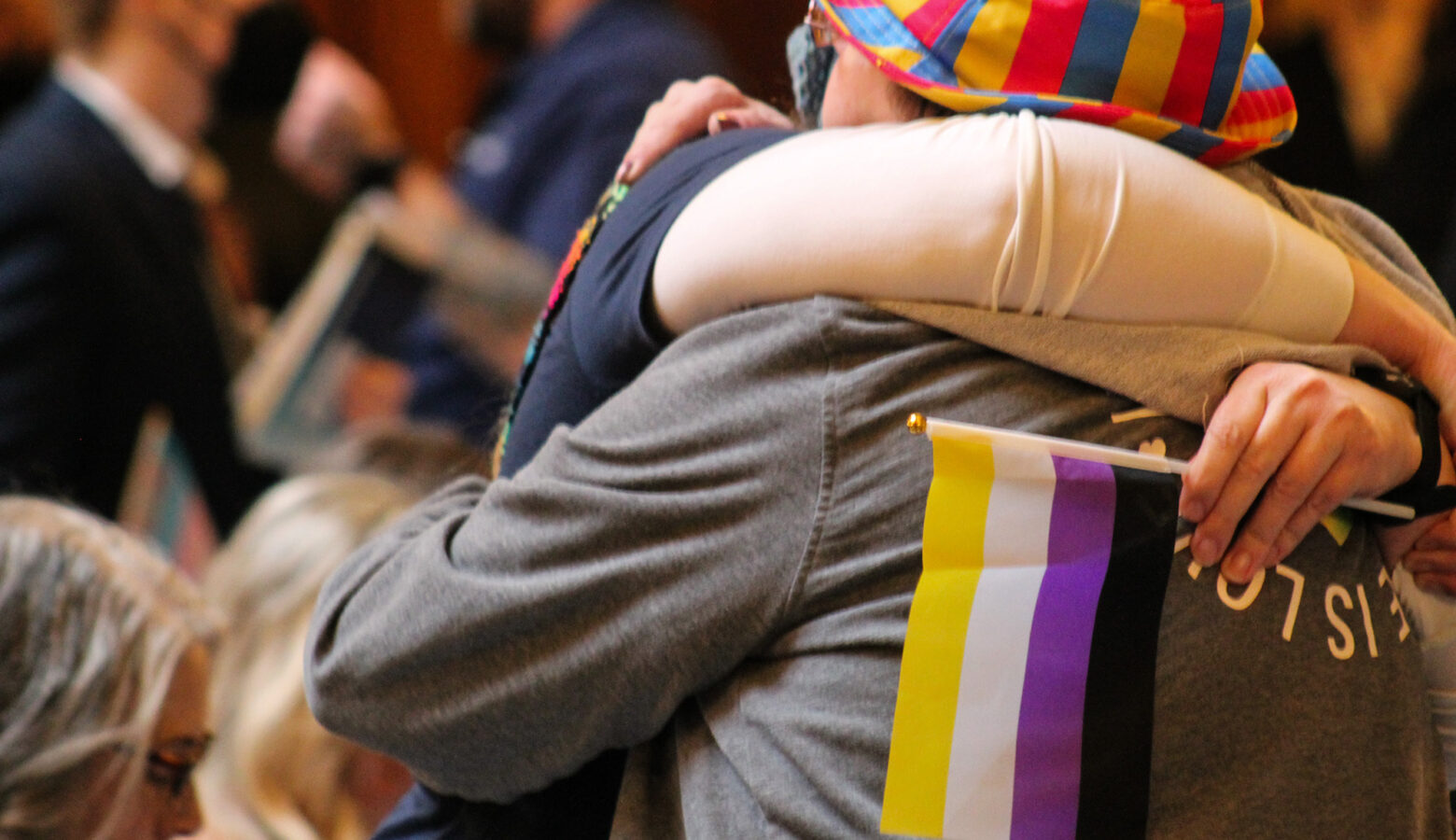 Two people embrace among a crowd inside the Indiana Statehouse. One person has a non-binary pride flag in their hand. The other wears a bucket hat with the pansexual pride colors.