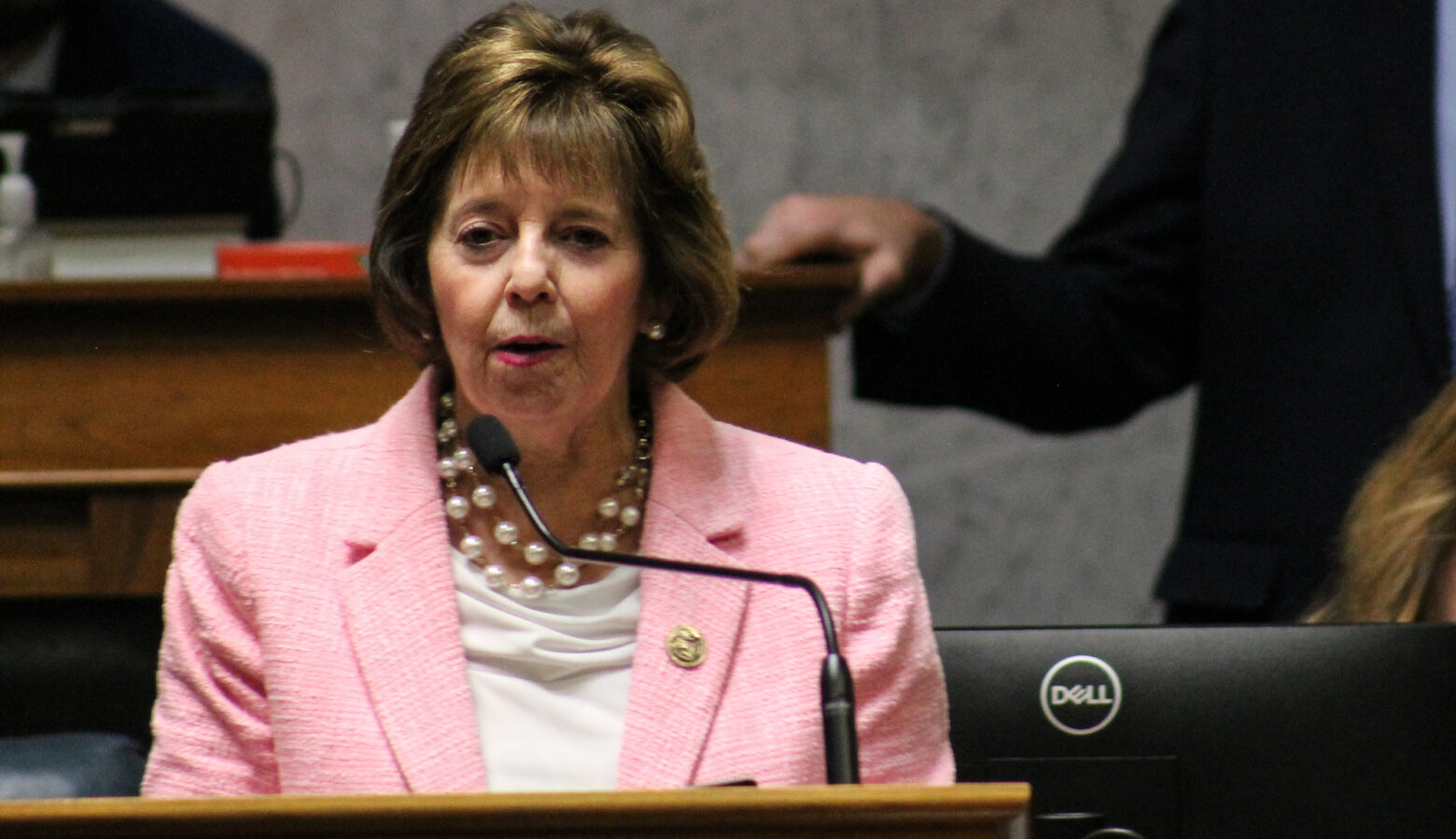 A woman in a pink suit jacket speaks into a microphone.