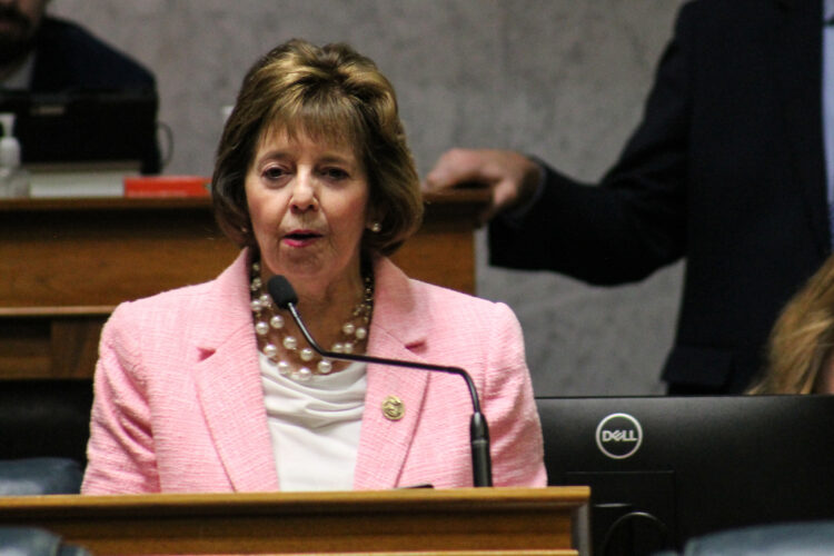 A woman in a pink suit jacket speaks into a microphone.