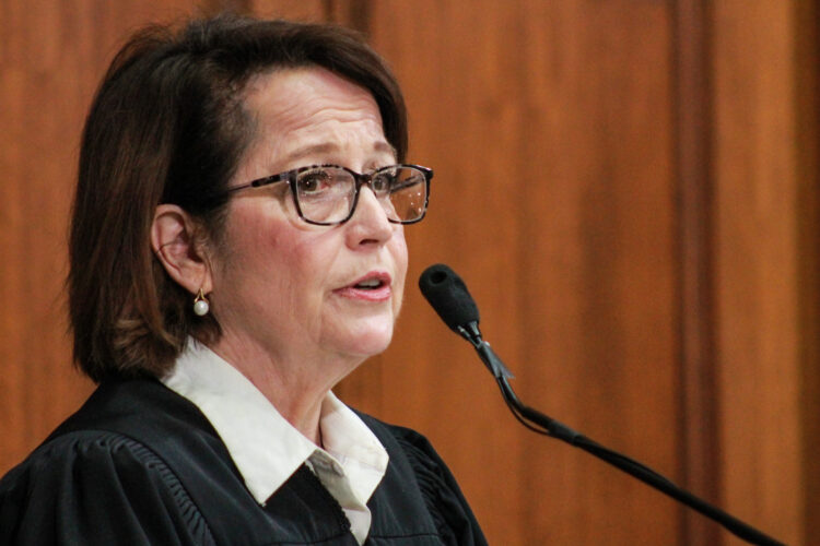 Loretta Rush is a White woman with dark brown hair. She is wearing glasses and black judicial robes over a white shirt.