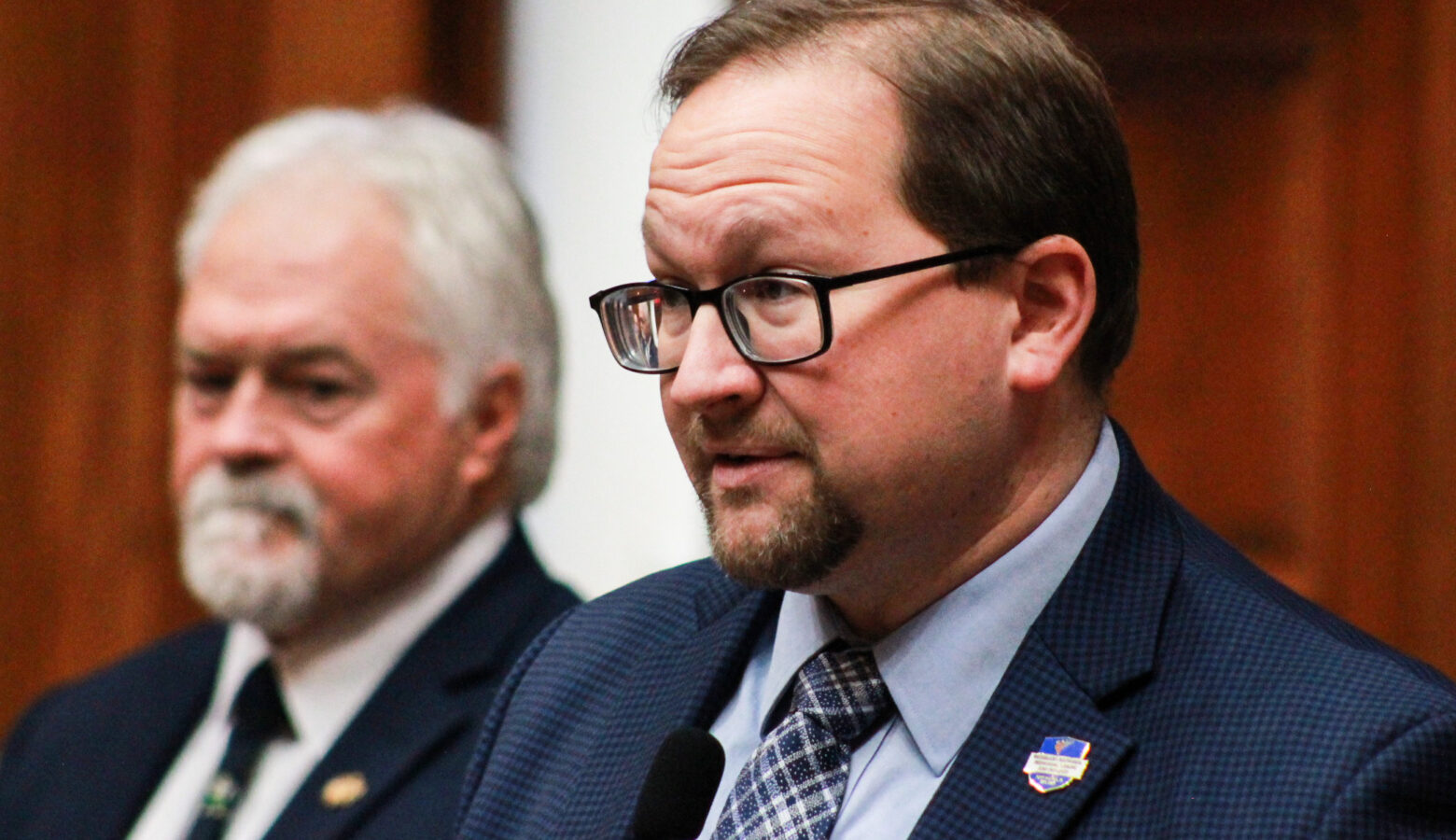 Martin Carbaugh is a White man with brown hair and goatee. He is wearing glasses and a blue suit. Brad Barrett is a White man with white hair and goatee. He is wearing a black suit.