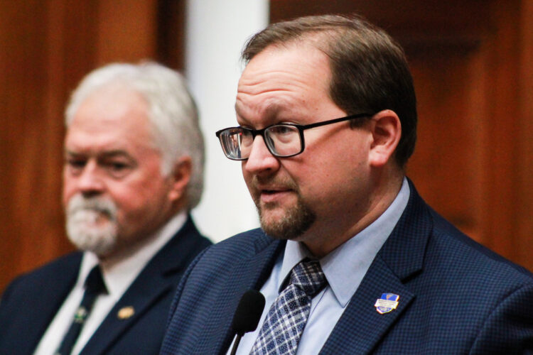 Martin Carbaugh is a White man with brown hair and goatee. He is wearing glasses and a blue suit. Brad Barrett is a White man with white hair and goatee. He is wearing a black suit.