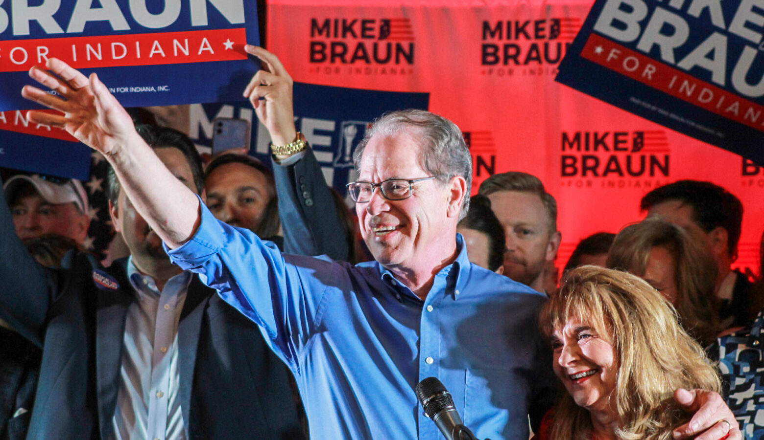 Mike Braun is a White man, balding with gray hair. He is wearing glasses and a blue shirt. Maureen is a White woman with dark blonde hair. She is wearing a red top.