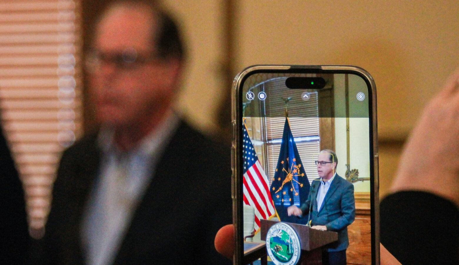 In the foreground, Mike Braun is seen on the screen of a cell phone that's taking a photo of him. In the background is Braun himself, out of focus. Braun is a White man with graying hair. He is wearing glasses and a dark blazer over a white shirt.