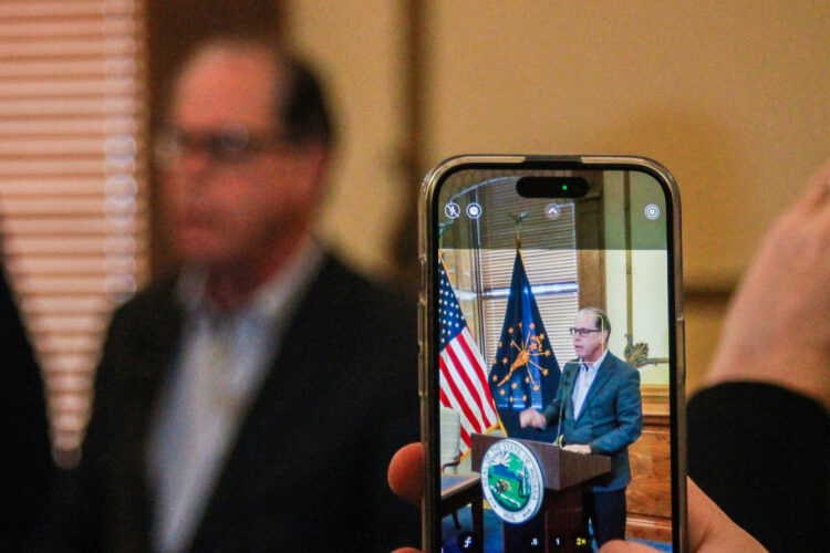 In the foreground, Mike Braun is seen on the screen of a cell phone that's taking a photo of him. In the background is Braun himself, out of focus. Braun is a White man with graying hair. He is wearing glasses and a dark blazer over a white shirt.