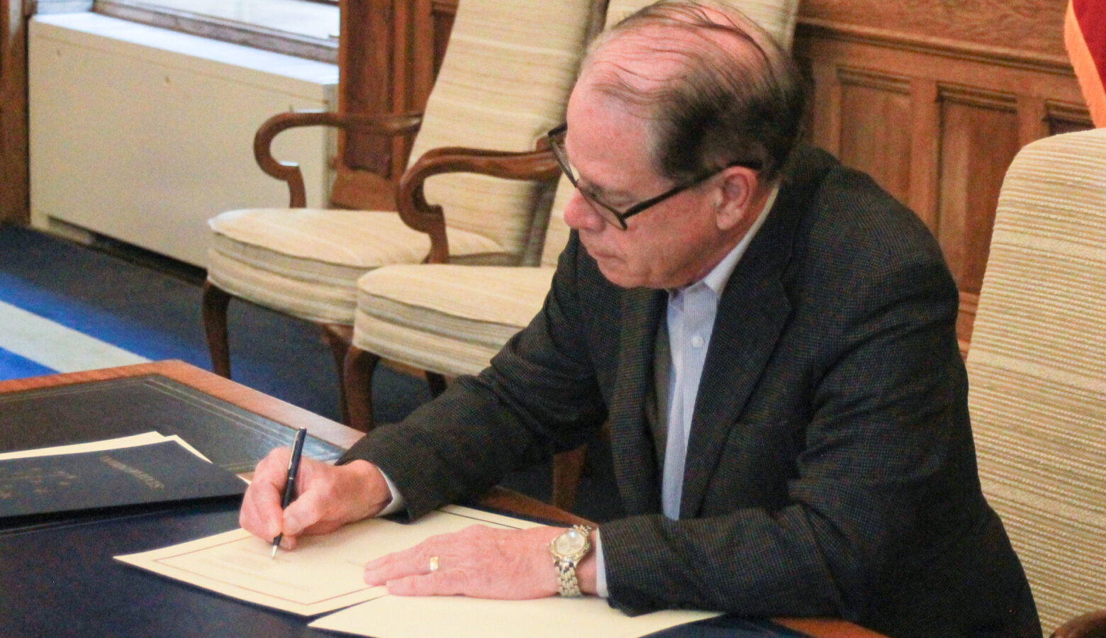 Mike Braun sits at a desk in his office, signing an executive order. Braun is a White man, balding with gray hair. He is wearing glasses and a dark blazer over a white shirt.