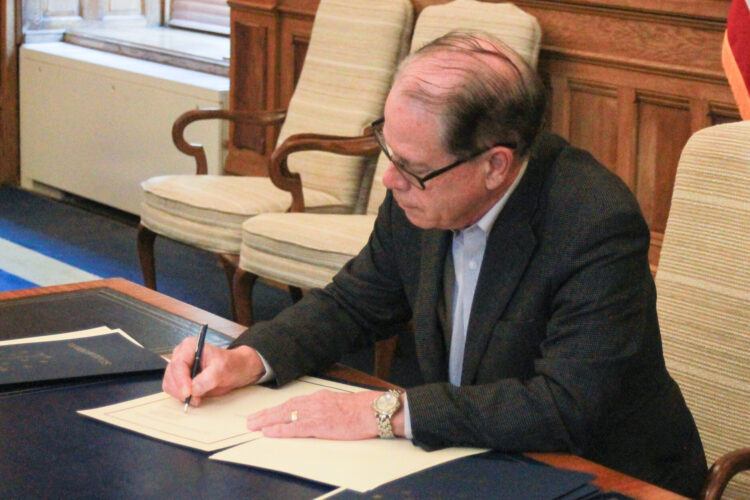 Mike Braun sits at a desk in his office, signing an executive order. Braun is a White man, balding with gray hair. He is wearing glasses and a dark blazer over a white shirt.