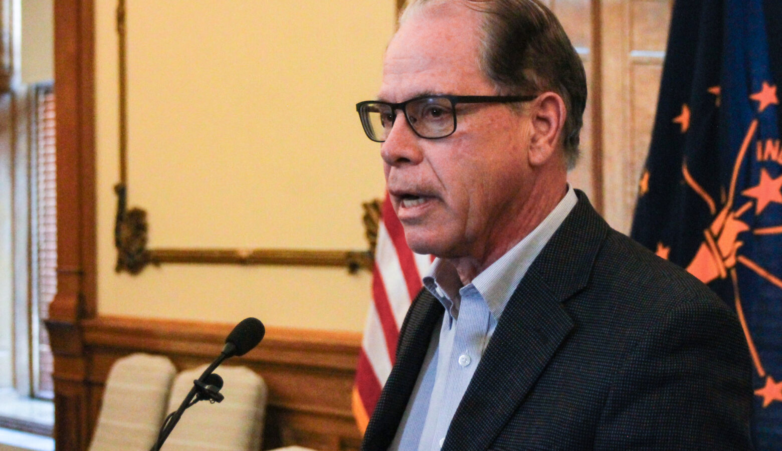 Mike Braun speaks into a microphone in his office. Braun is a White man, balding with gray hair. He is wearing glasses and a dark blazer over a white shirt.