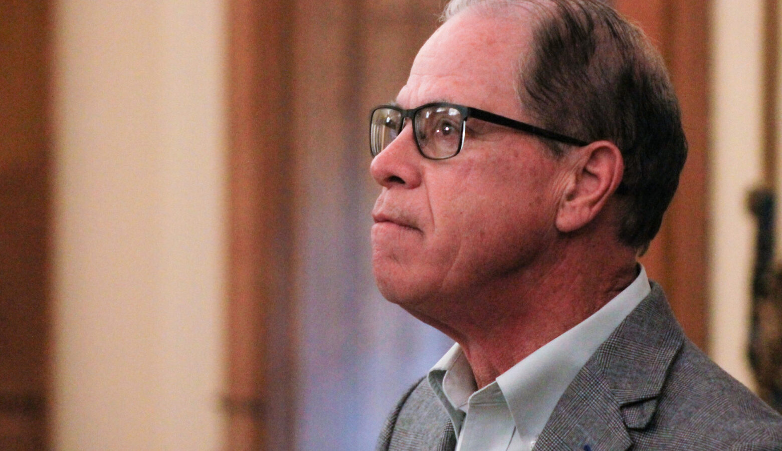 Mike Braun looks upward as he stands in his office. Braun is a White man, balding with gray hair. He is wearing glasses and a gray suit jacket over a white shirt