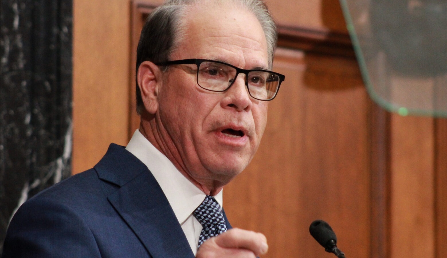 Mike Braun gestures while speaking in the Indiana House. Braun is a White man, balding with gray hair. He is wearing glasses and a blue suit.