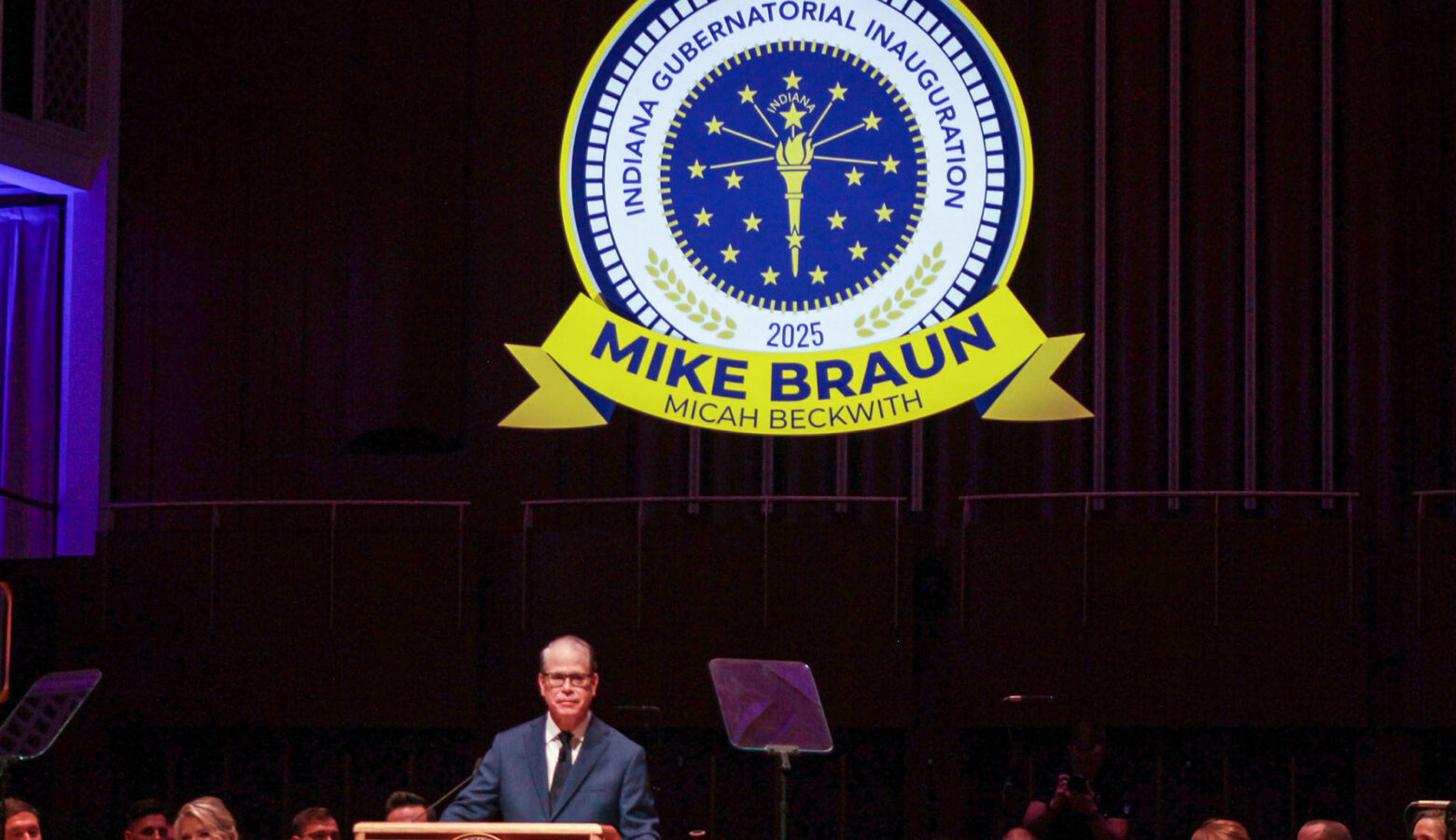 Mike Braun stands on stage at a lectern while a logo for the inauguration ceremony hangs above him. Braun is a White man, balding with gray hair. He is wearing glasses and a suit.