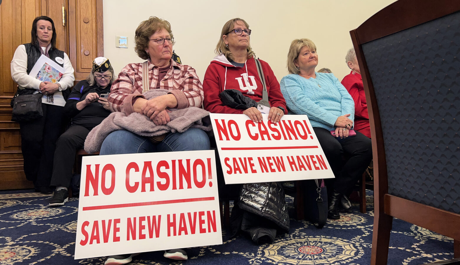 Two women sit in a committee room, each with a sign in front of them that reads "No Casino! Save New Haven"