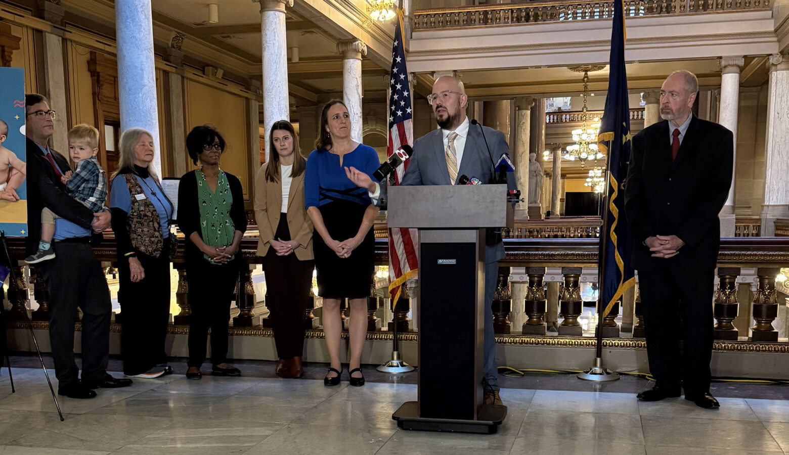 There is a man wearing a grey blazer and white dress shirt while he is standing at a podium. There are people behind him standing beside each other looking at him speak.