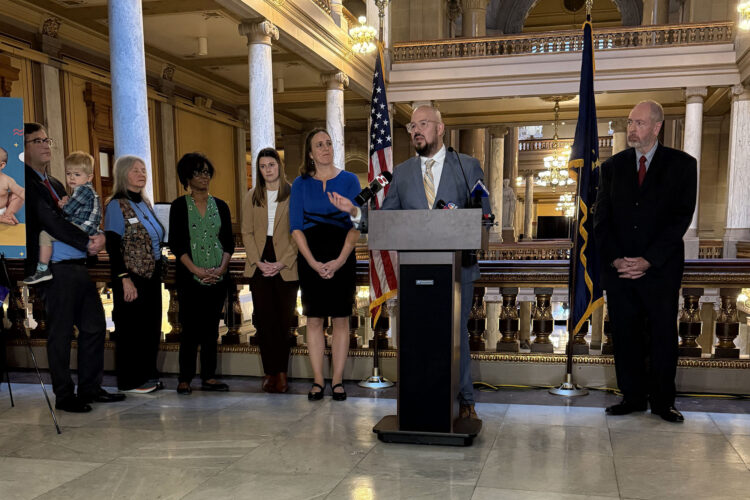 There is a man wearing a grey blazer and white dress shirt while he is standing at a podium. There are people behind him standing beside each other looking at him speak.