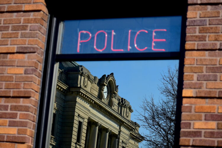 A neon sign in a window reads "Police." There is a county courthouse in the reflection of the window.