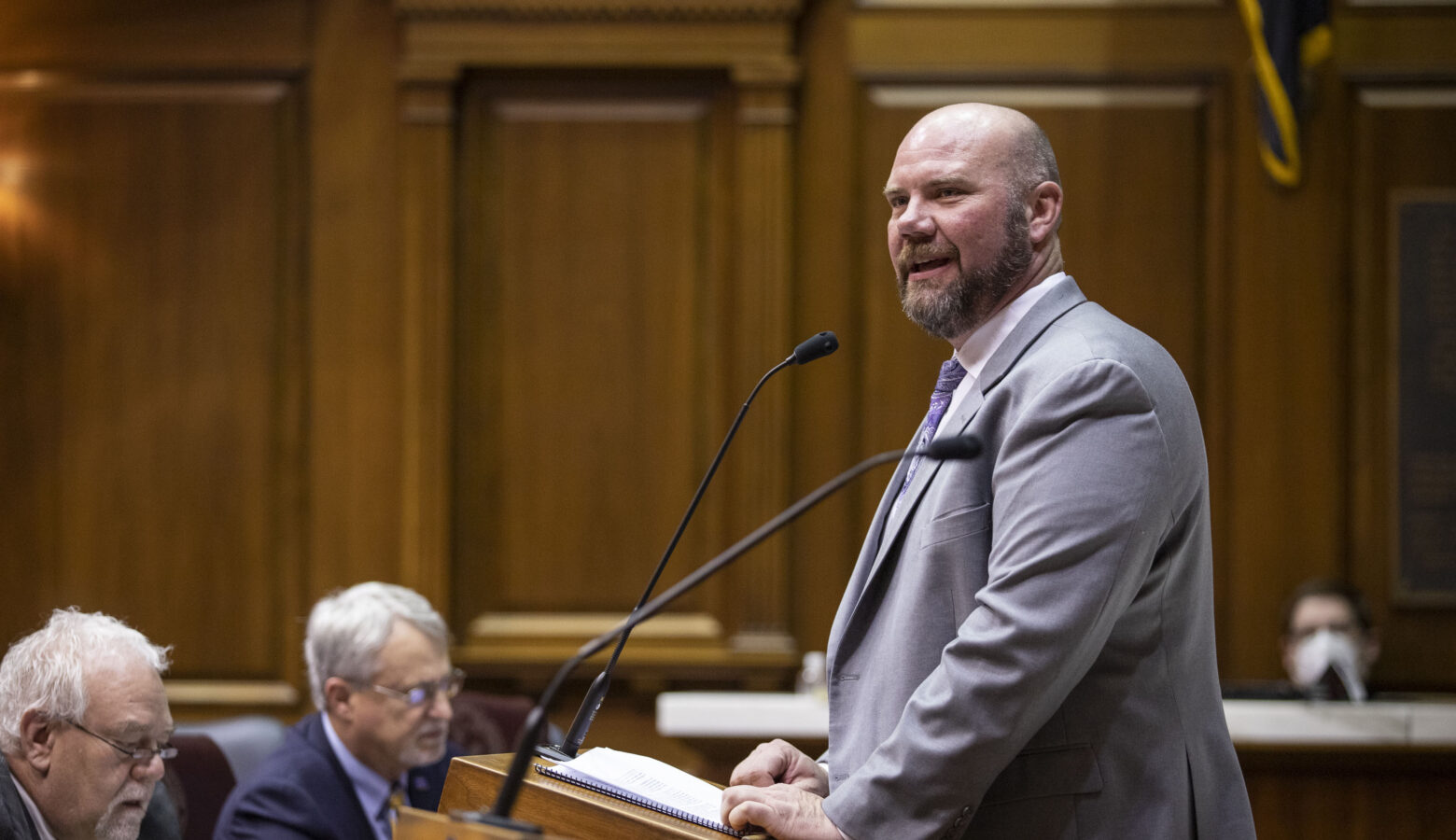 Alan Morrison speaks on the House floor at a podium. He is a White man with a beard, wearing a grey suit.