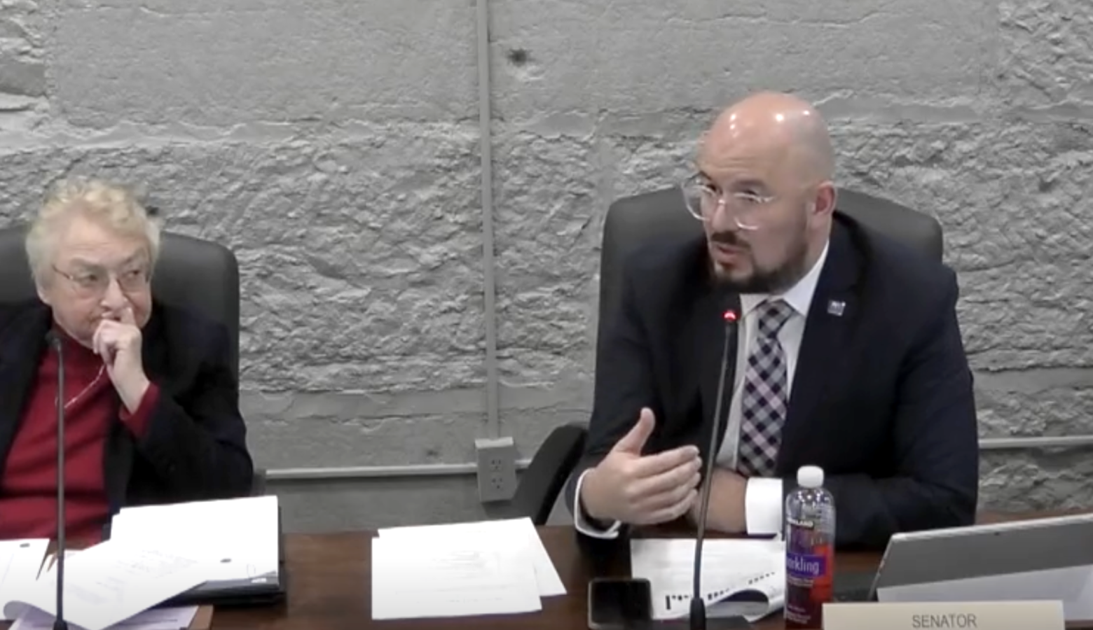 Rodney Pol sits at a desk and speaks into a microphone while a Sue Glick watches him.