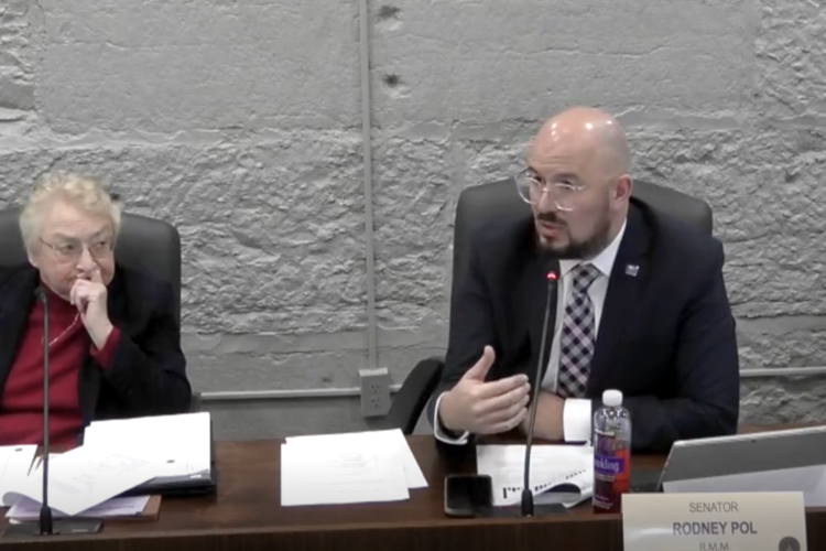 Rodney Pol sits at a desk and speaks into a microphone while a Sue Glick watches him.