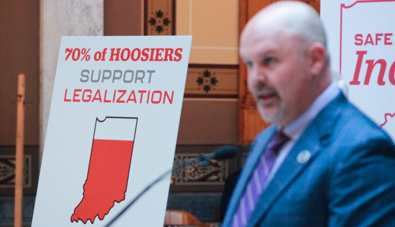 A poster on an easel reads "70 percent of Hoosiers support legalization." Heath VanNatter is in the foreground of the image, out of focus.