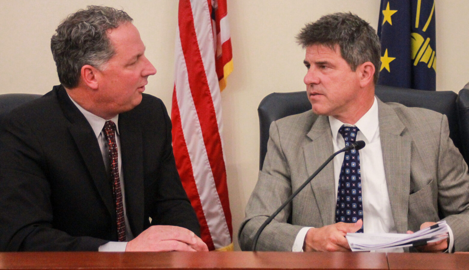 Todd Huston and Rodric Bray sit next to each other, speaking to each other. Huston is a White man with dark, graying hair. He is wearing a black suit. Bray is a White man with dark, graying hair. He is wearing a gray suit.