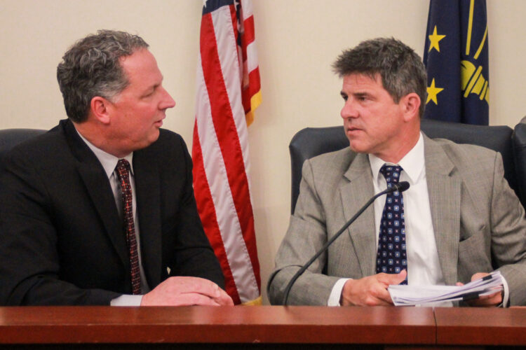 Todd Huston and Rodric Bray sit next to each other, speaking to each other. Huston is a White man with dark, graying hair. He is wearing a black suit. Bray is a White man with dark, graying hair. He is wearing a gray suit.