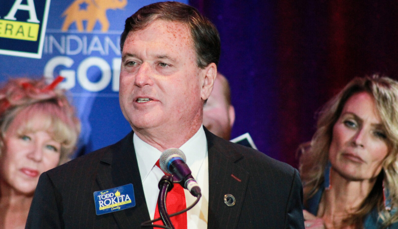 Todd Rokita speaks at a campaign event. Rokita is a White man with dark hair. He is wearing a black suit, white shirt and red tie.