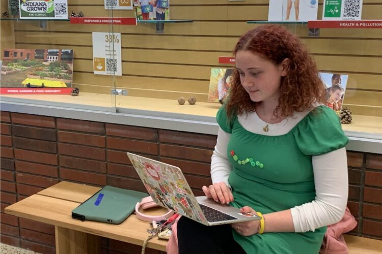 A White student with red curly hair sits on a bench, typing on a laptop in her lap.