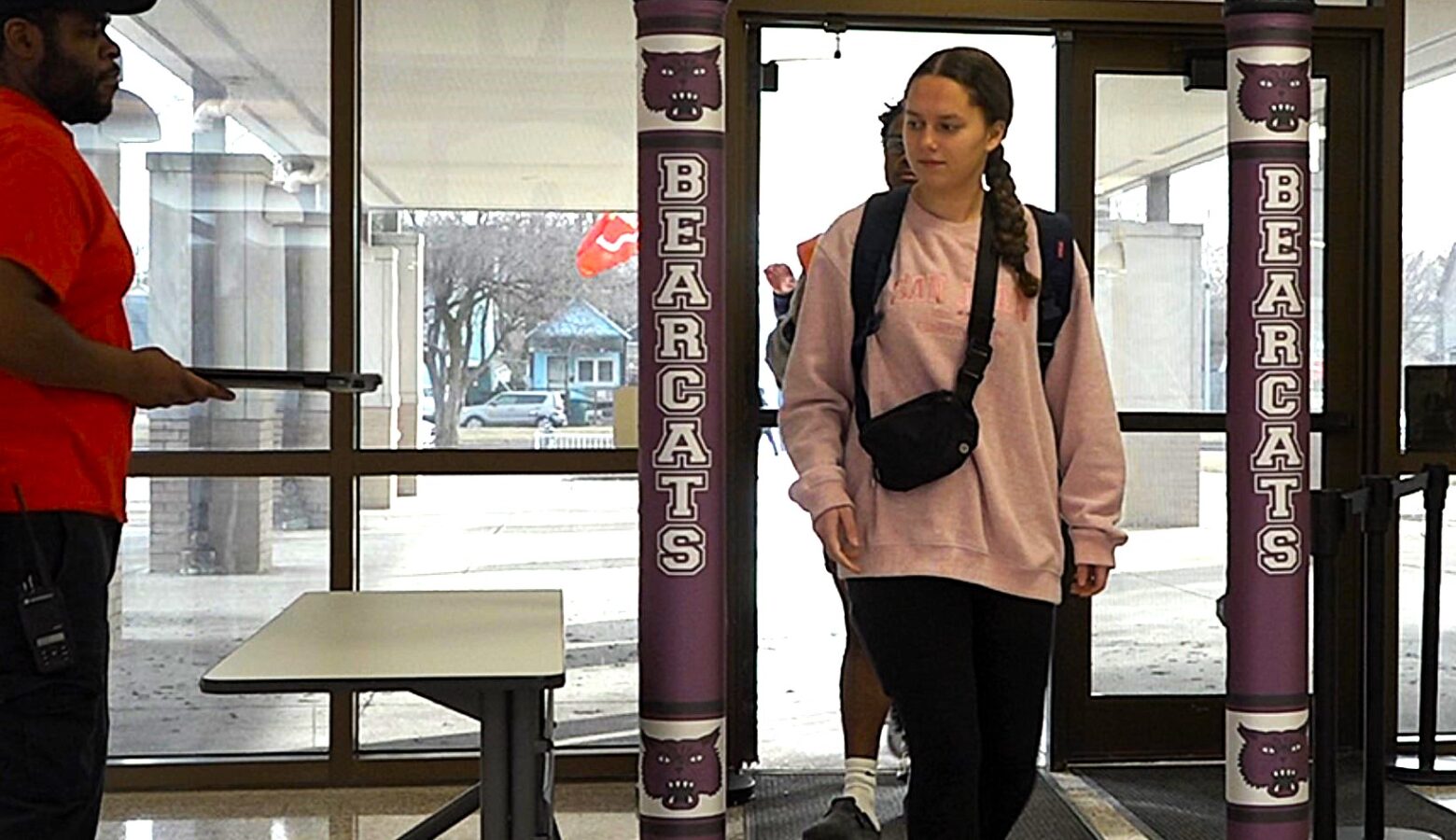 A student in a pink sweater and carrying a black bag walks through a door between two purple pillars of a security scanner.