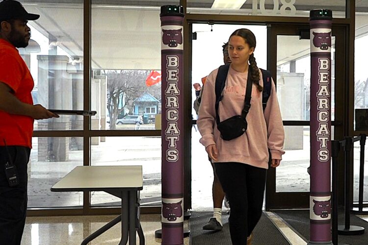 A student in a pink sweater and carrying a black bag walks through a door between two purple pillars of a security scanner.