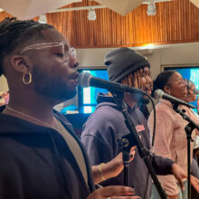 Four casually dressed people smile and sing into microphones in front of windows in a performance studio.