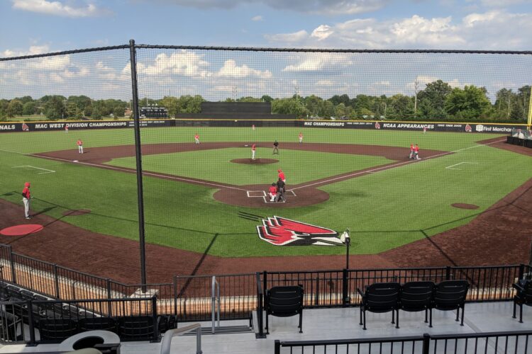 From the stands and behind a black fence, baseball players in red and white uniforms play on a baseball field.