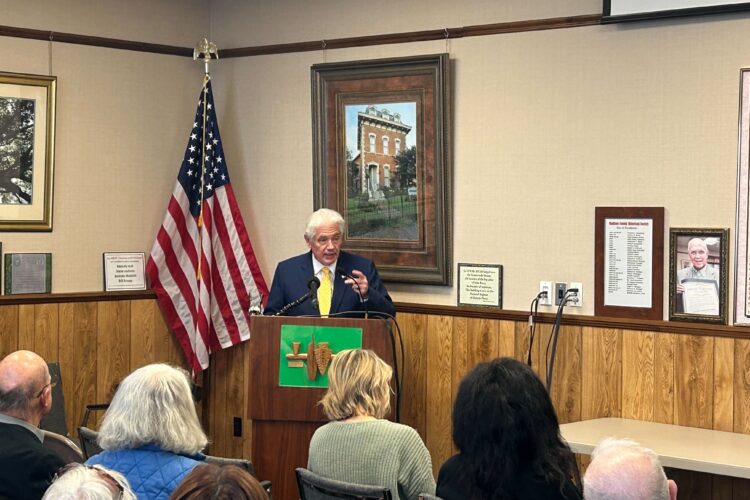 A man in a suit stands at a podium addressing a crowd in front of him.