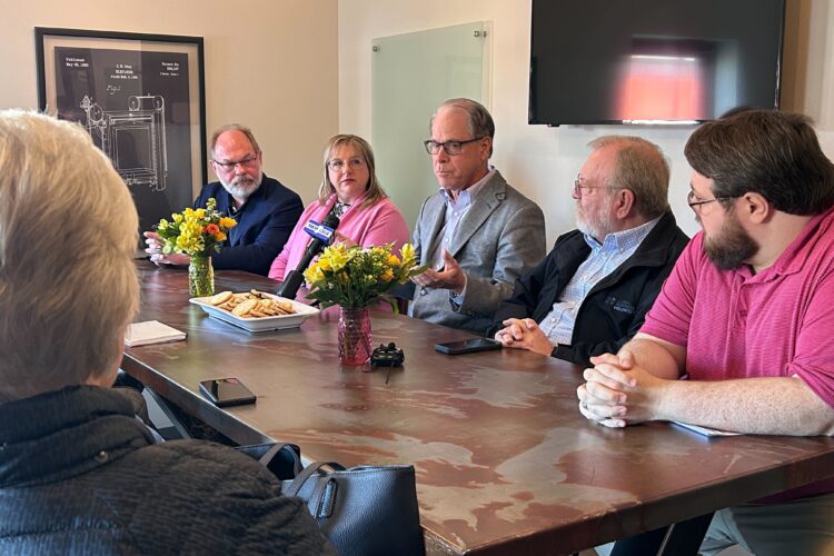 Gov. Mike Braun, an older White man in a suit, sits around a table with people listening to him speak.