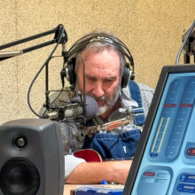 A man with salt-and-pepper hair and a striped shirt looks down in thought. He's wearing headphones, and we see him between a speaker, a microphone, and other audio equiptment.