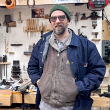 A smiling man in a denim overshirt and safety glasses stands in front of a messy workbench with tools and materials.