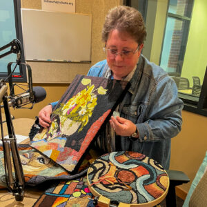 A woman in a denim shirt holds several framed rugs. The rugs are cut and dyed to look like paintings.