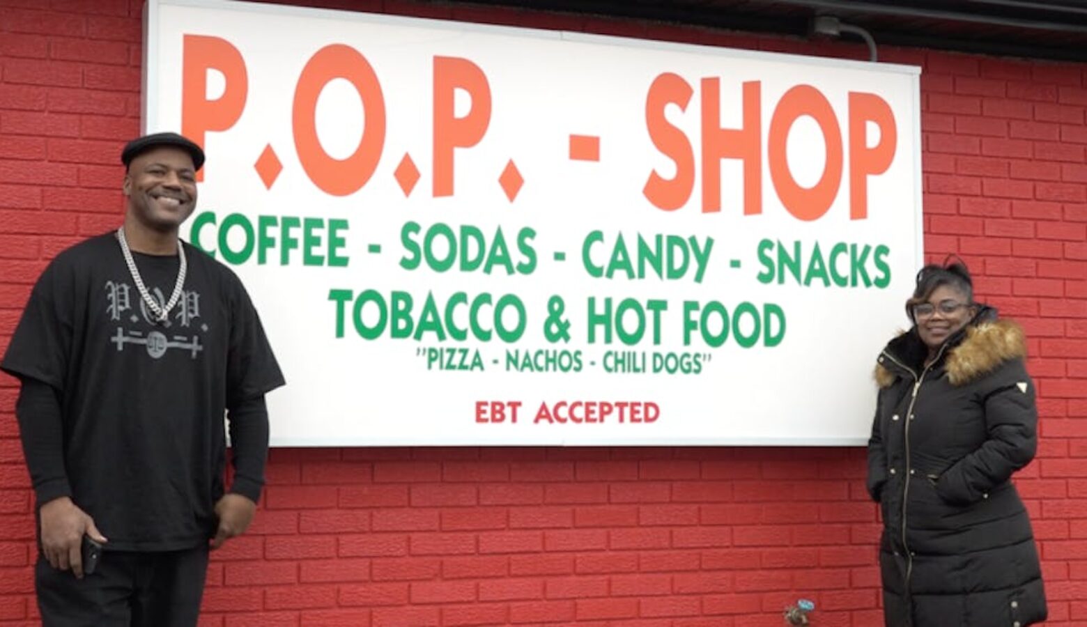 Paris Powell and his fiancé stand in front of a red painted building with the shop's sign between them.