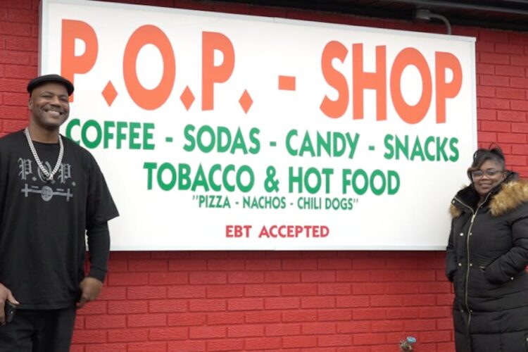 Paris Powell and his fiancé stand in front of a red painted building with the shop's sign between them.