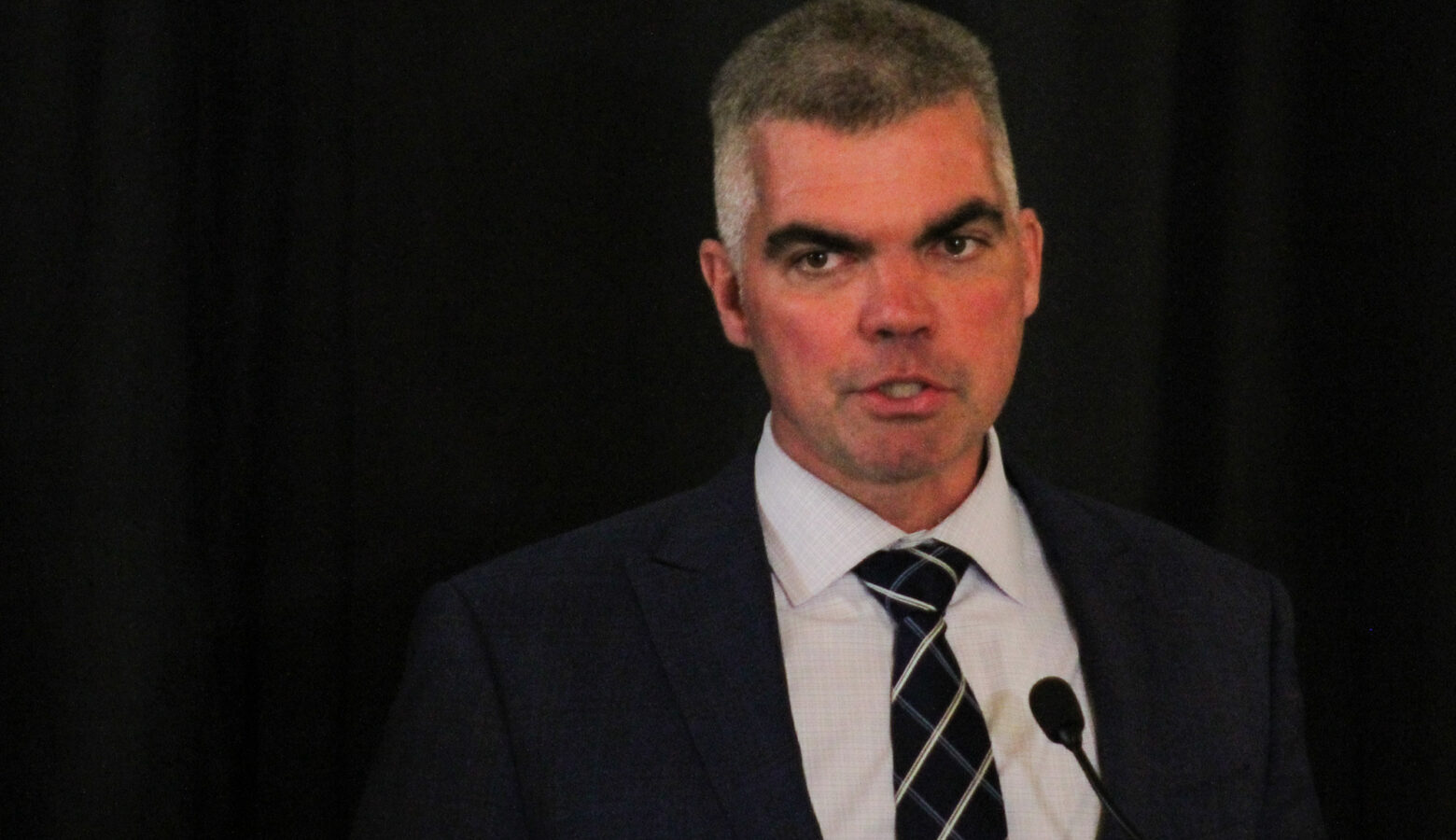 Bob Morris stands in front of a dark blue curtain, wearing a suit and tie. He is a White man with a grey buzzcut that is white at the temples.